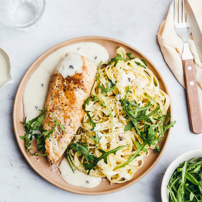 A beautifully plated dish of pan-seared chicken breast and tagliatelle pasta is served with a creamy white sauce and garnished with fresh arugula and black pepper.