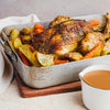 A golden-brown roasted chicken, seasoned with herbs, rests in a metal pan with roasted potatoes, carrots, and fennel, with a side of gravy in a white boat.