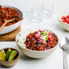A hearty bowl of chili con carne is served over white rice and garnished with sour cream, diced onions, tomatoes, and fresh jalapeños.