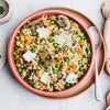 A pink bowl filled with a creamy grain dish, possibly risotto, mixed with sliced mushrooms, green peas, and pieces of chicken, garnished with shaved Parmesan cheese and fresh herbs.