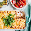 A freshly baked lasagna in a white baking dish is served with a side of tomato salad and garlic bread.