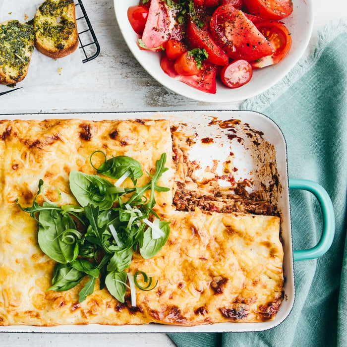 A freshly baked lasagna in a white baking dish is served with a side of tomato salad and garlic bread.