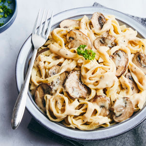 A close-up shot shows a delicious-looking bowl of creamy mushroom fettuccine, garnished with fresh parsley and black pepper, with a fork resting on the side of the bowl.