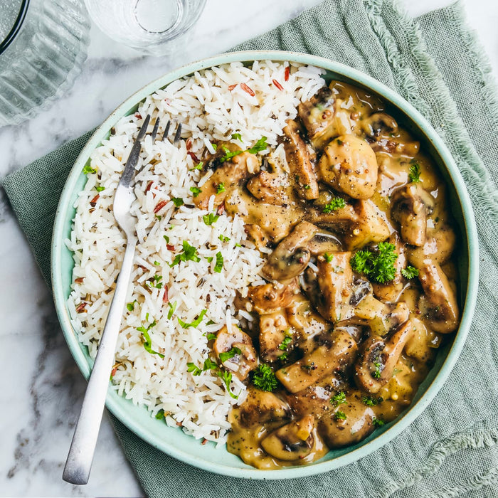 A delicious-looking bowl of creamy mushroom stew is served alongside wild rice and garnished with fresh parsley and black pepper.