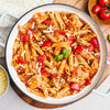 A top-down view of a white pot filled with a hearty chicken and red pepper penne pasta in a tomato sauce, garnished with shredded cheese and fresh basil.