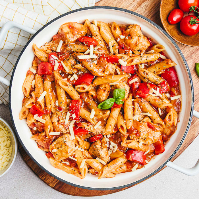 A top-down view of a white pot filled with a hearty chicken and red pepper penne pasta in a tomato sauce, garnished with shredded cheese and fresh basil.