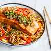 A glazed salmon fillet rests on a bed of noodles with red and yellow peppers, garnished with sliced green chilies and sesame seeds in a bowl.