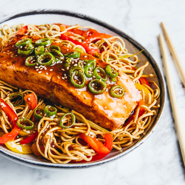 A glazed salmon fillet rests on a bed of noodles with red and yellow peppers, garnished with sliced green chilies and sesame seeds in a bowl.