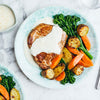A seared pork chop is plated with roasted potatoes, carrots, and broccolini, all drizzled with a creamy white sauce, on a marble countertop.