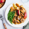 A white plate holds a serving of pork and apple stew with roasted potatoes and green beans, viewed from above.
