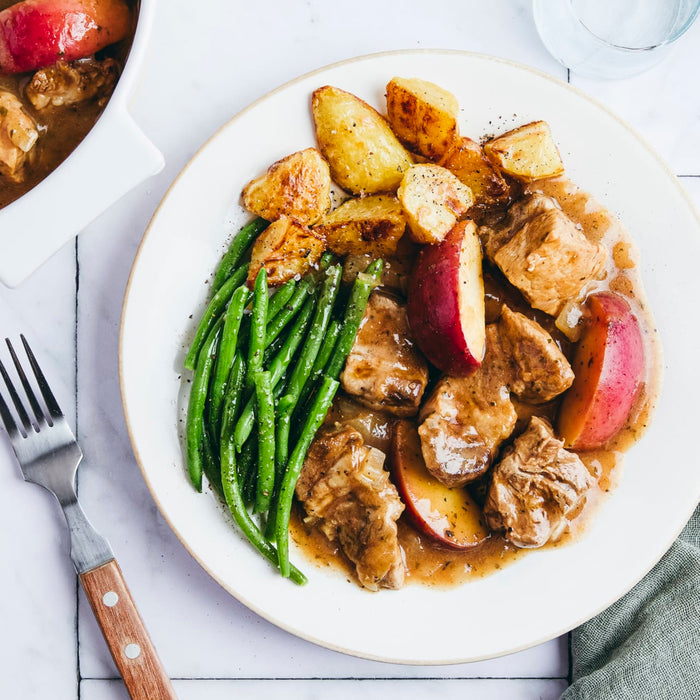 A white plate holds a serving of pork and apple stew with roasted potatoes and green beans, viewed from above.