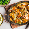 A hearty meal is presented from a top-down perspective, featuring a rustic pan of braised chicken thighs with carrots and mushrooms, alongside bowls of mashed potatoes and green beans.