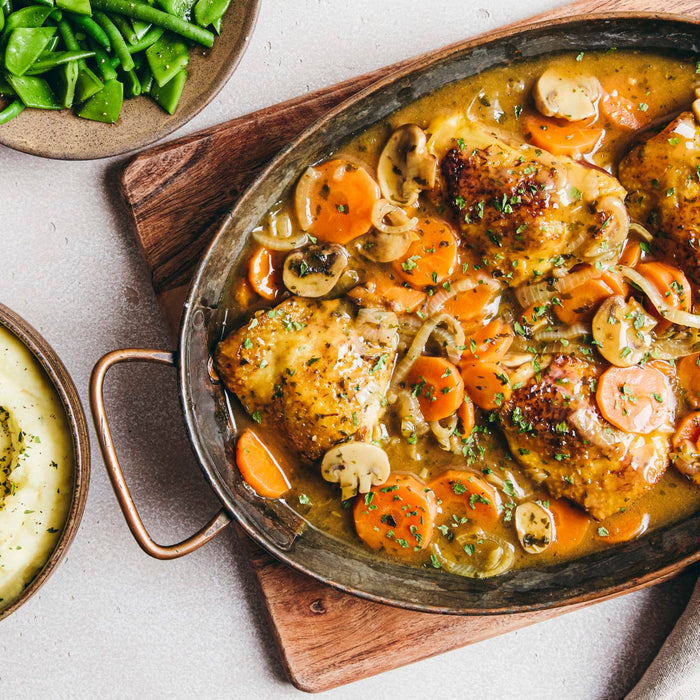 A hearty meal is presented from a top-down perspective, featuring a rustic pan of braised chicken thighs with carrots and mushrooms, alongside bowls of mashed potatoes and green beans.