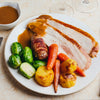 A plated roast turkey dinner with gravy, roasted potatoes, carrots, Brussels sprouts, and a bacon-wrapped sausage.
