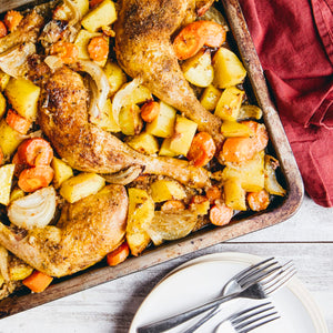 A rustic sheet pan meal featuring golden-brown roasted chicken legs and a medley of chopped potatoes, carrots, and onions, set on a white wooden table.