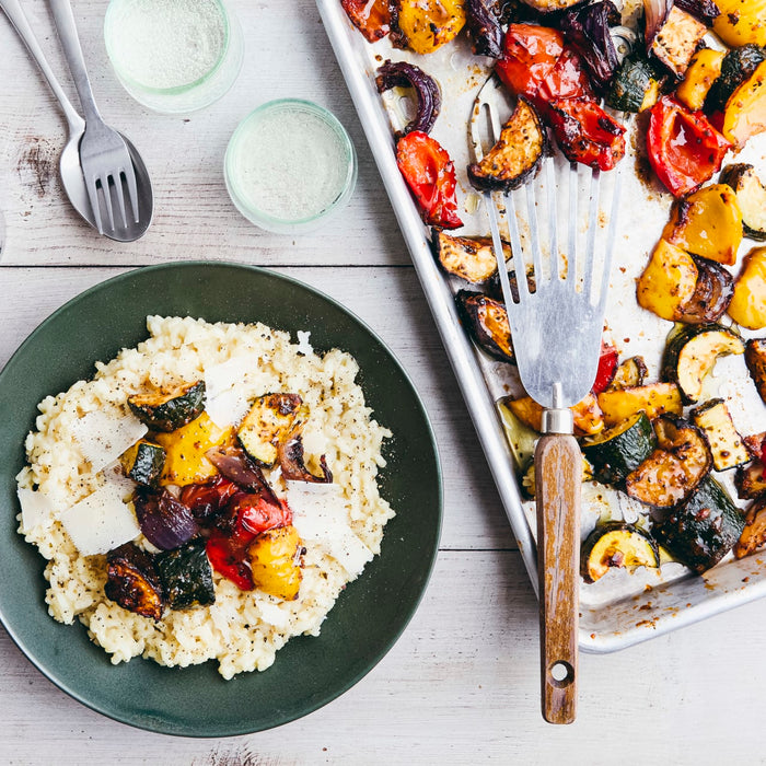 A dark green plate holds a serving of creamy risotto topped with seasoned roasted vegetables and shaved Parmesan cheese, with a baking sheet of more vegetables nearby.