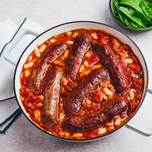 A white enamel pot holds a hearty sausage and bean casserole in a rich tomato sauce, served with a side of fresh snow peas.