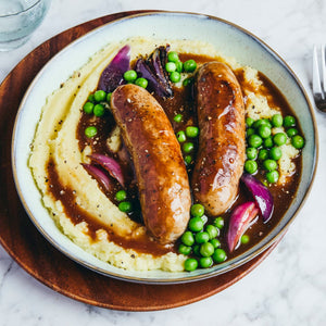 A hearty bowl of bangers and mash features two sausages nestled in creamy mashed potatoes with peas, red onions, and a rich gravy, all seasoned with a sprinkle of black pepper.