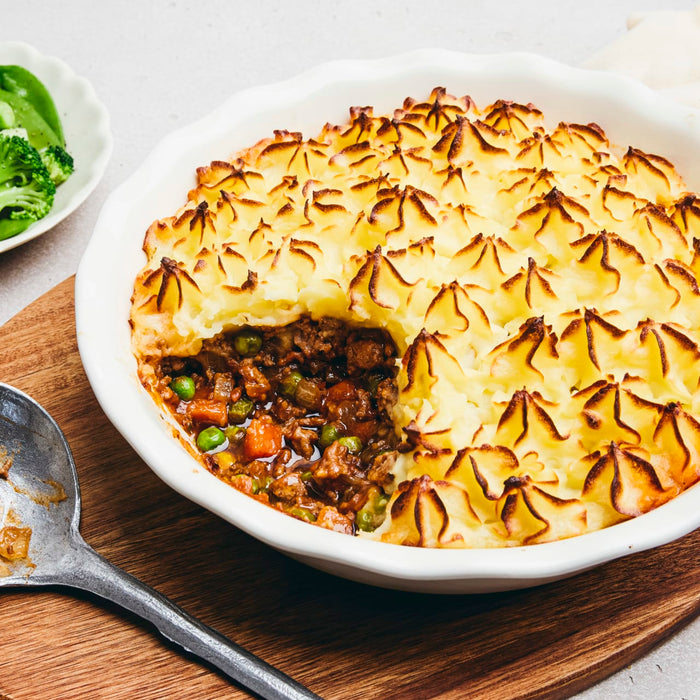 A freshly baked shepherd's pie in a white dish has a scoop taken out, revealing the savory meat and vegetable filling beneath the golden-brown piped potato topping.