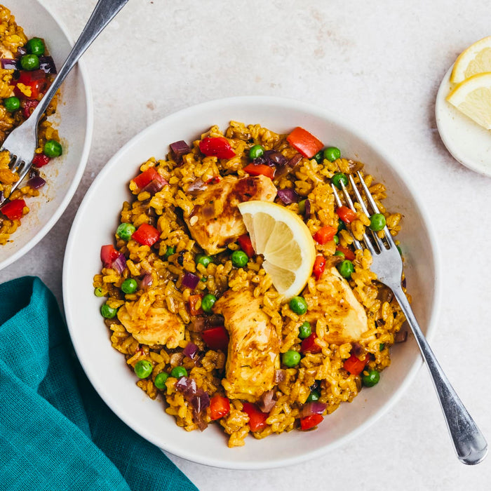 Two white bowls are filled with a savory chicken and rice dish, mixed with peas and red peppers, and garnished with a lemon wedge.