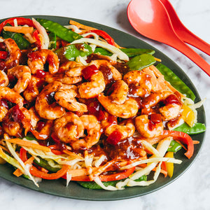 A close-up, overhead shot of a platter of shrimp stir-fry with snow peas, shredded carrots, and bell peppers, served on a marble surface with red spoons.