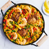 A vibrant paella is served in a traditional pan, featuring yellow rice, chicken, shrimp, chorizo, peas, and peppers, with a side of lemon wedges.