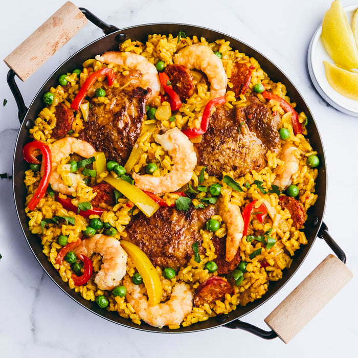 A vibrant paella is served in a traditional pan, featuring yellow rice, chicken, shrimp, chorizo, peas, and peppers, with a side of lemon wedges.