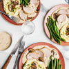 An overhead shot shows several plates of sliced pork tenderloin with a creamy mushroom sauce, served alongside mashed potatoes and green beans on a white marble table.