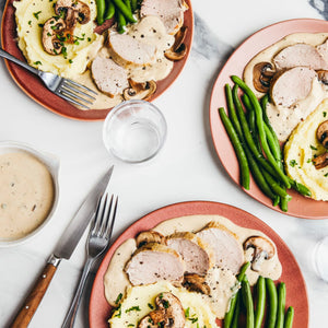 An overhead shot shows several plates of sliced pork tenderloin with a creamy mushroom sauce, served alongside mashed potatoes and green beans on a white marble table.