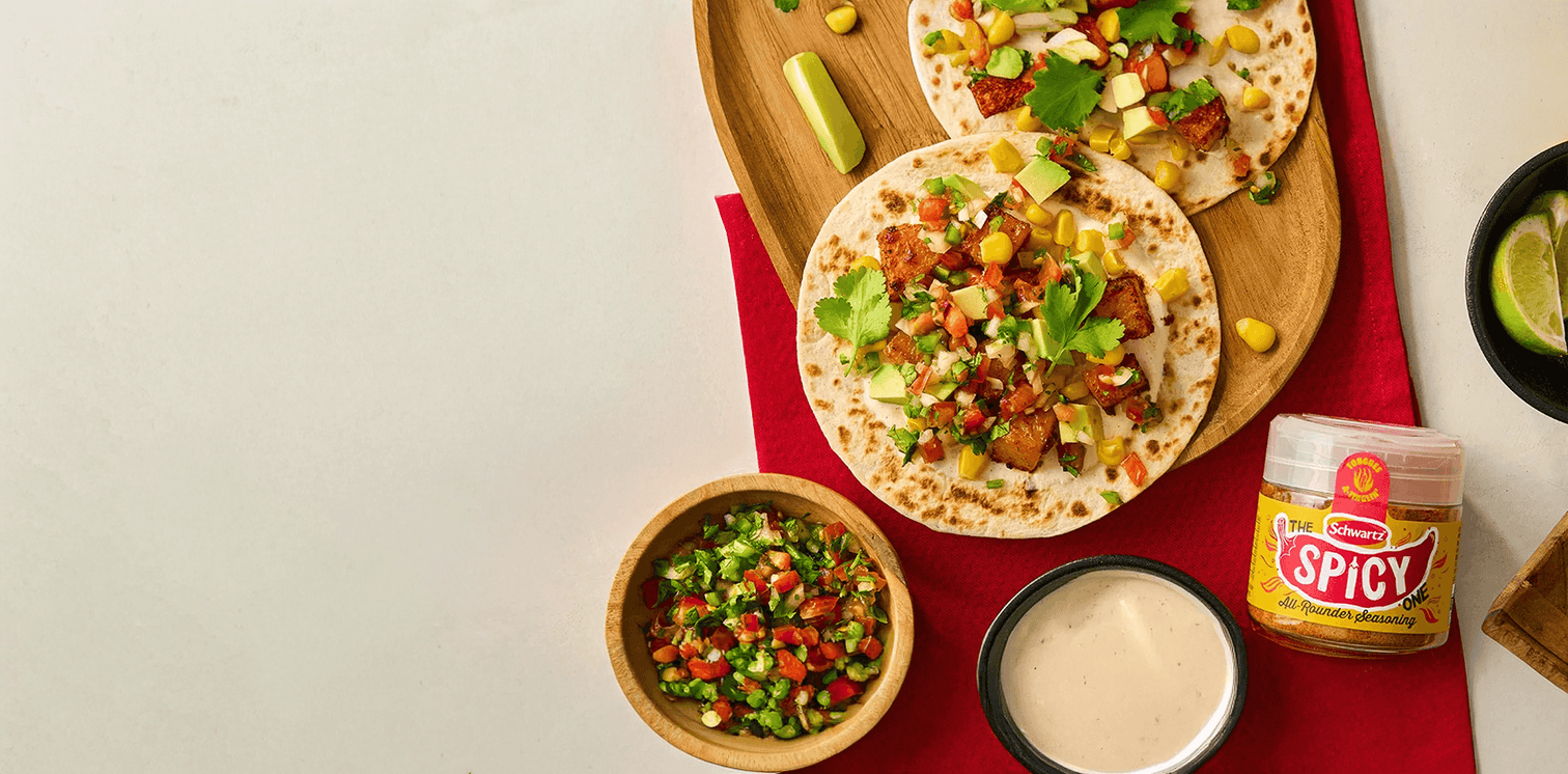 Tortillas with toppings on a wooden board, accompanied by a bowl of salad, sour cream, and a jar of spicy all rounder seasoning. 
