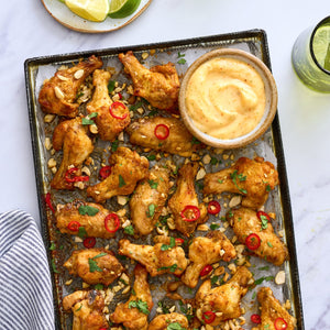 A baking sheet is filled with golden-brown chicken wings, garnished with peanuts, red chili, and cilantro, and served with a creamy dipping sauce.