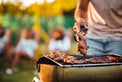 Person grilling meat on a barbecue with people in the background