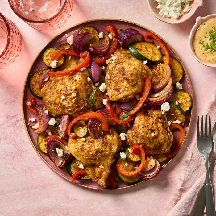 A platter of roasted chicken thighs and vegetables, seasoned with a Barrel spice blend, is garnished with feta cheese and served with a creamy dipping sauce.