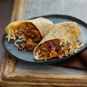 Two savory pork and coleslaw wraps are served on a rustic blue plate, which is placed on a wooden Barrel surface.