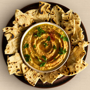 A top-down view of a bowl of hummus, swirled with olive oil and garnished with fresh parsley and a sprinkle of paprika, surrounded by pieces of toasted pita bread on a dark platter.