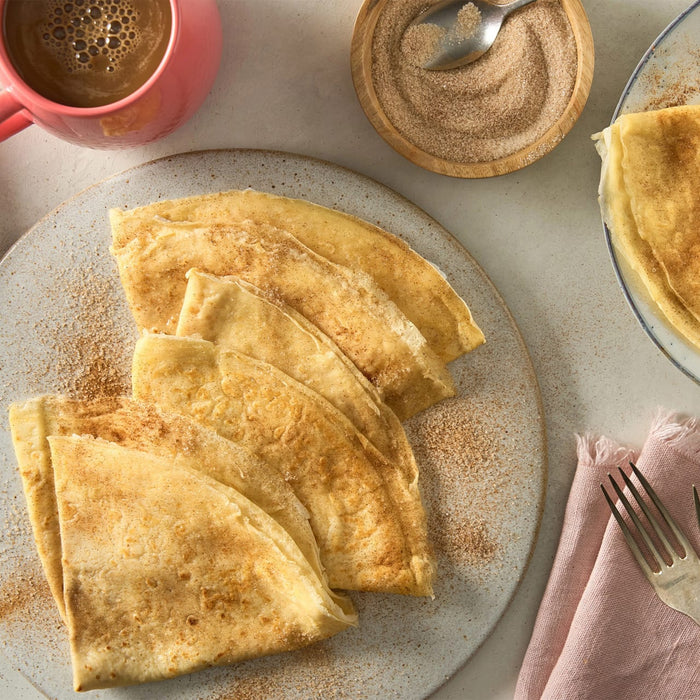 Folded crepes are sprinkled with a cinnamon-sugar mixture from a small wooden Barrel, served on a plate next to a cup of coffee.