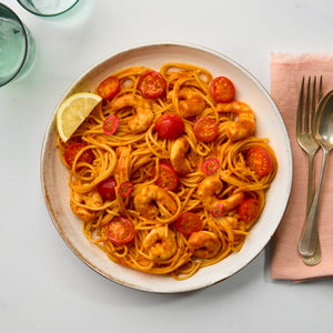 An overhead shot shows a serving of shrimp and tomato spaghetti in a rustic, speckled Barrel bowl, garnished with a lemon wedge and sliced red chili.
