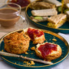 An afternoon tea is served on dark green plates from the Barrel collection, featuring a savory scone split and topped with butter and jam, with finger sandwiches and a cup of tea in the background.