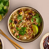 A bowl of Asian noodle soup is seen from above, garnished with fresh cilantro, green onions, a lime wedge, and chili oil, with chopsticks and side dishes nearby.
