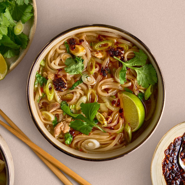 A bowl of Asian noodle soup is seen from above, garnished with fresh cilantro, green onions, a lime wedge, and chili oil, with chopsticks and side dishes nearby.