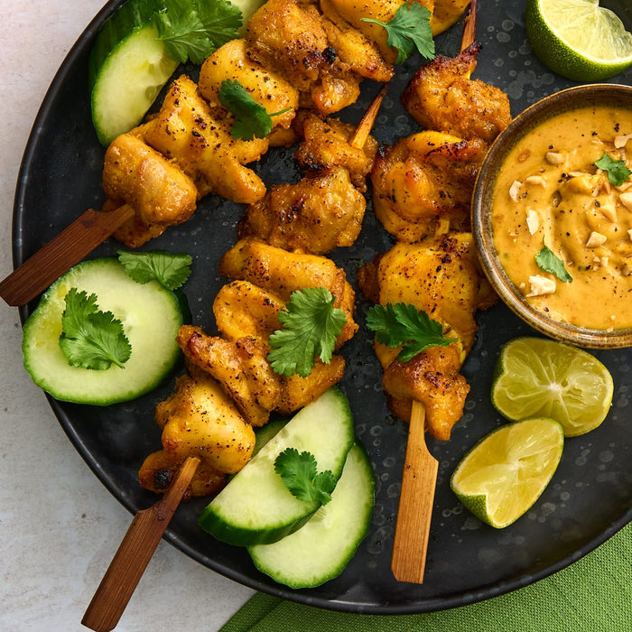 An overhead view of chicken satay skewers on a dark platter, served with a creamy peanut sauce in a small Barrel bowl, and garnished with fresh cucumber, lime, and cilantro.