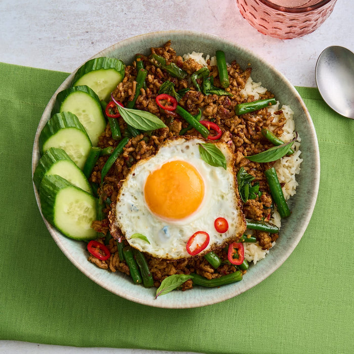 An overhead view of a Thai basil stir-fry with minced meat, green beans, and rice, served in a rustic Barrel bowl and topped with a fried egg and fresh cucumber slices.