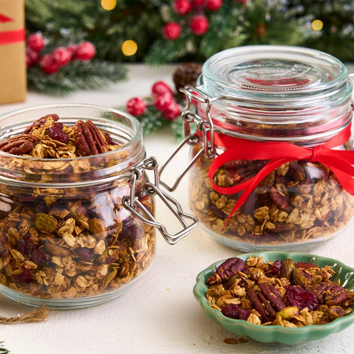 Homemade Christmas granola with pecans and cranberries is shown in two glass Barrel jars and a small green bowl, set against a festive background.