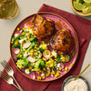 Two glazed and roasted chicken thighs are served on a pink plate alongside a fresh salad with corn, avocado, radishes, and feta cheese.