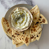 A creamy dip topped with dill and olive oil is presented in a rustic Barrel bowl, served with toasted pita bread on a marble surface.