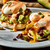 A close-up, appetizing shot of Barrel chorizo eggs benedict, featuring poached eggs with runny yolks over avocado and English muffins, with a bottle of Cholula hot sauce in the background.