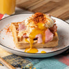 A savory waffle topped with ham and a perfectly poached egg is seasoned with a generous sprinkle of a spice blend like the Barrel seasoning, with the golden yolk running down the side.