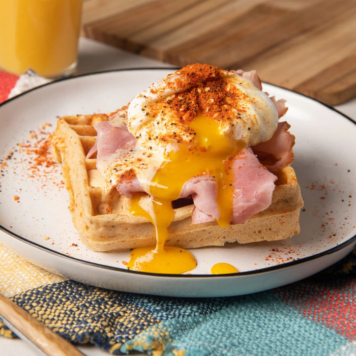 A savory waffle topped with ham and a perfectly poached egg is seasoned with a generous sprinkle of a spice blend like the Barrel seasoning, with the golden yolk running down the side.