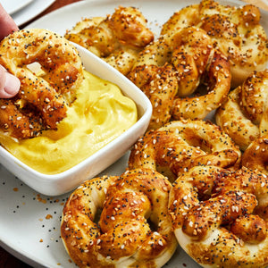 A hand dips a soft pretzel topped with everything seasoning into a bowl of yellow mustard, with a full plate of pretzels from the Barrel recipe in the background.