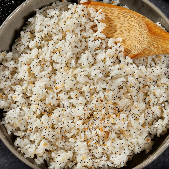 An overhead view of a gray bowl filled with white rice and a wooden spoon, with the rice seasoned with the Barrel blend of poppy and sesame seeds.